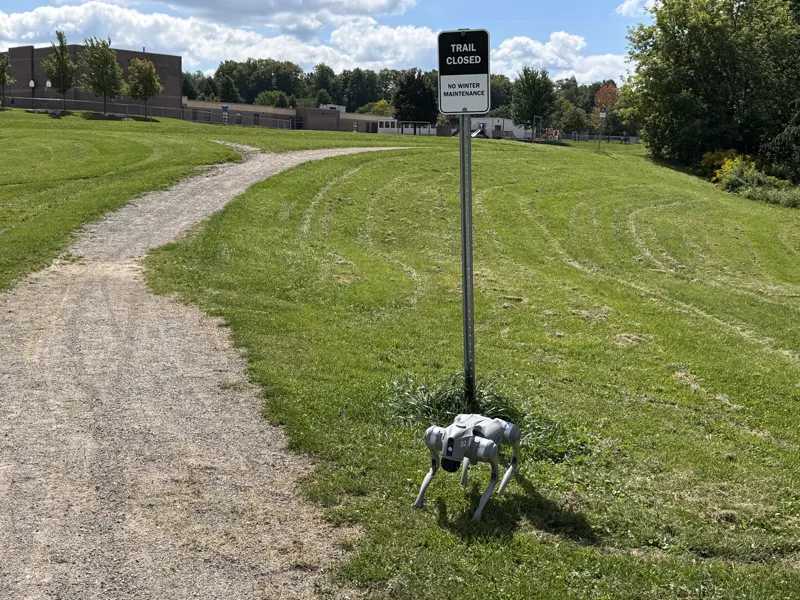 A grey Go2 robot dog on grass beside a Trail Closed sign